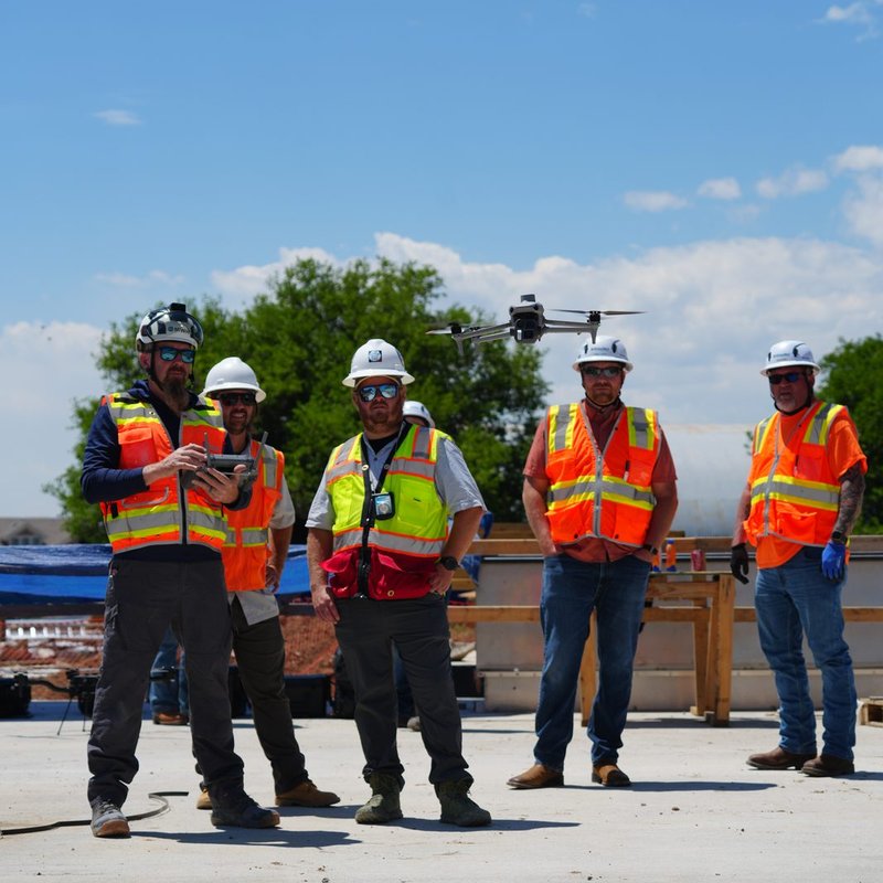 Drone U crew of six in hard hats and hi-vis vests coordinating as the pilot flies above a landing pad, the teamwork behind scaling a drone program