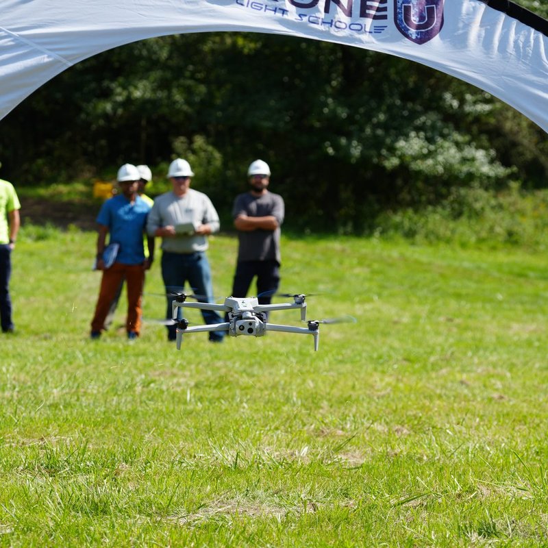 Close-up of a drone hovering through a Drone U Flight School gate while trainees in hard hats observe