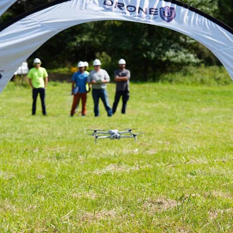 View through a Drone U Flight School gate of a drone hovering low with trainees in hard hats standing behind it