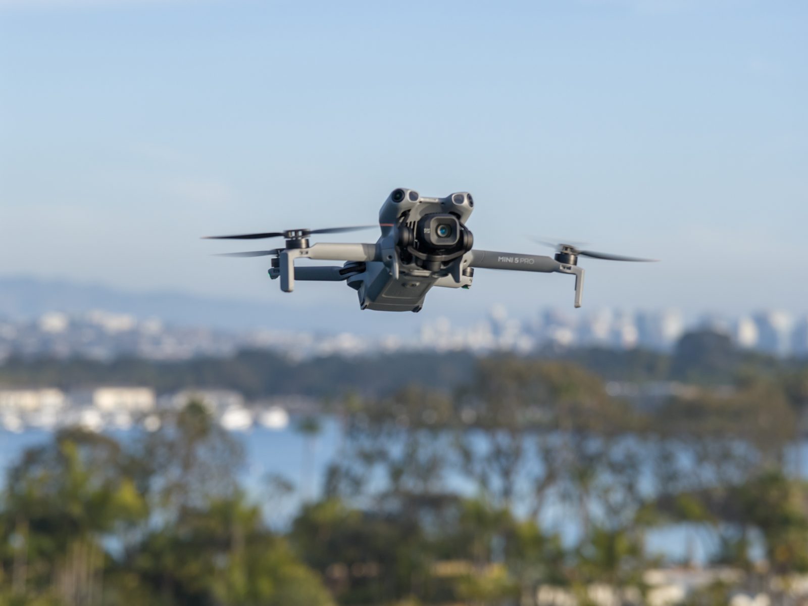 DJI Mini 5 Pro drone in flight with a city skyline and bay in the background